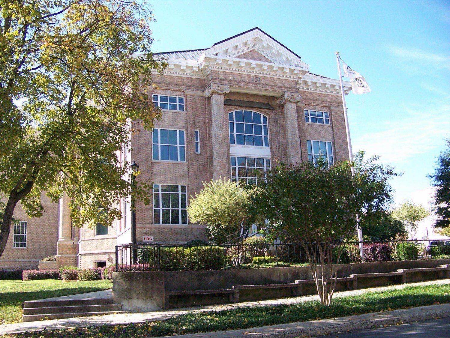 Neoclassical tan brick courthouse with columns and a pedimented entrance, shaded by trees, in downtown Gastonia, North Carolina