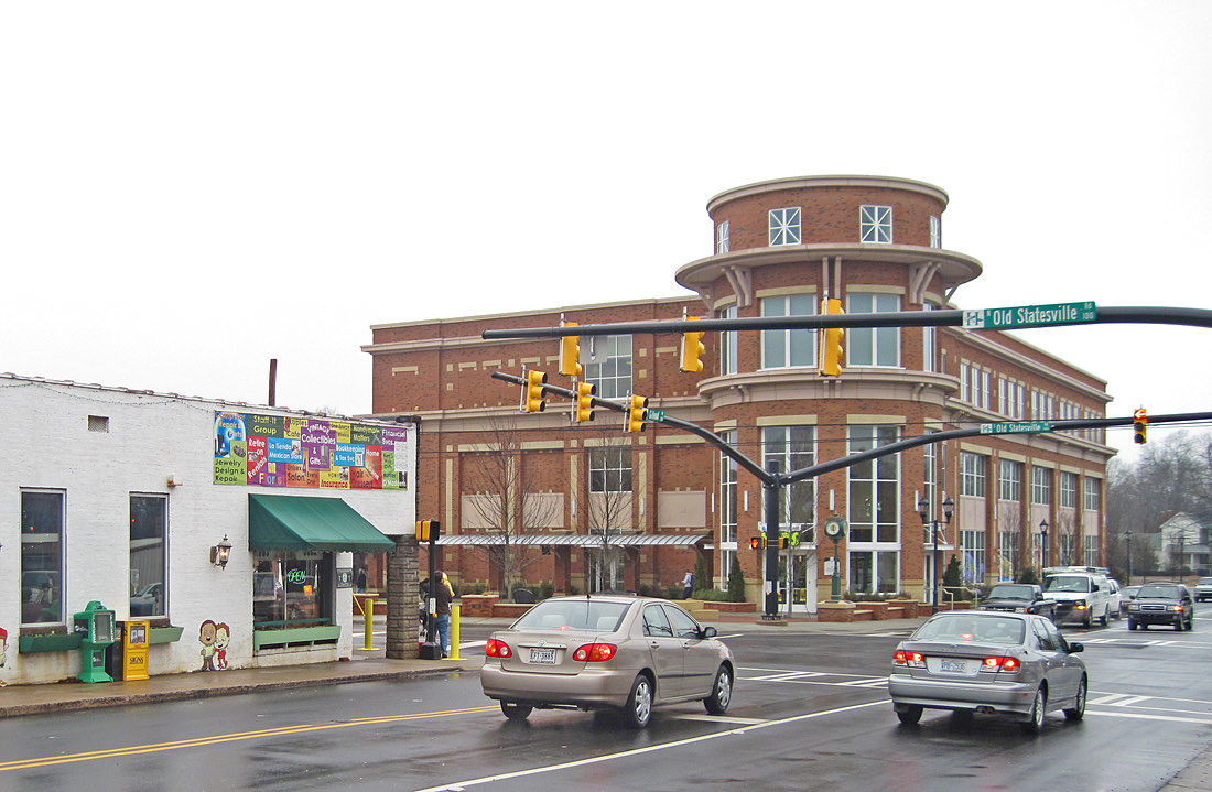 Street corner in downtown Huntersville, North Carolina, with a rounded brick and stucco office building, a small local shop, and Old Statesville Road street signs above the intersection