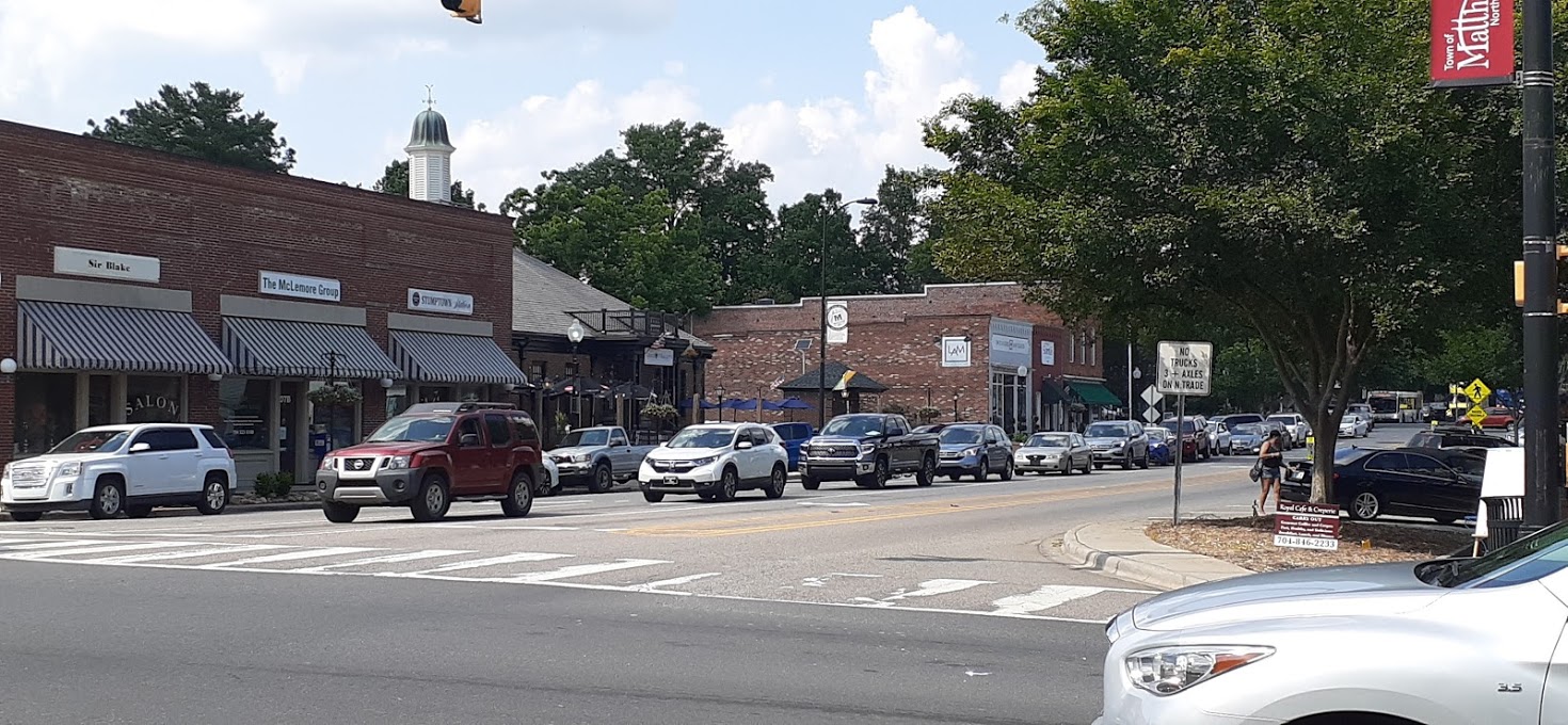 Awning-covered shopfronts and a red Town of Matthews street banner along North Trade Street in downtown Matthews, North Carolina, with traffic passing through the intersection