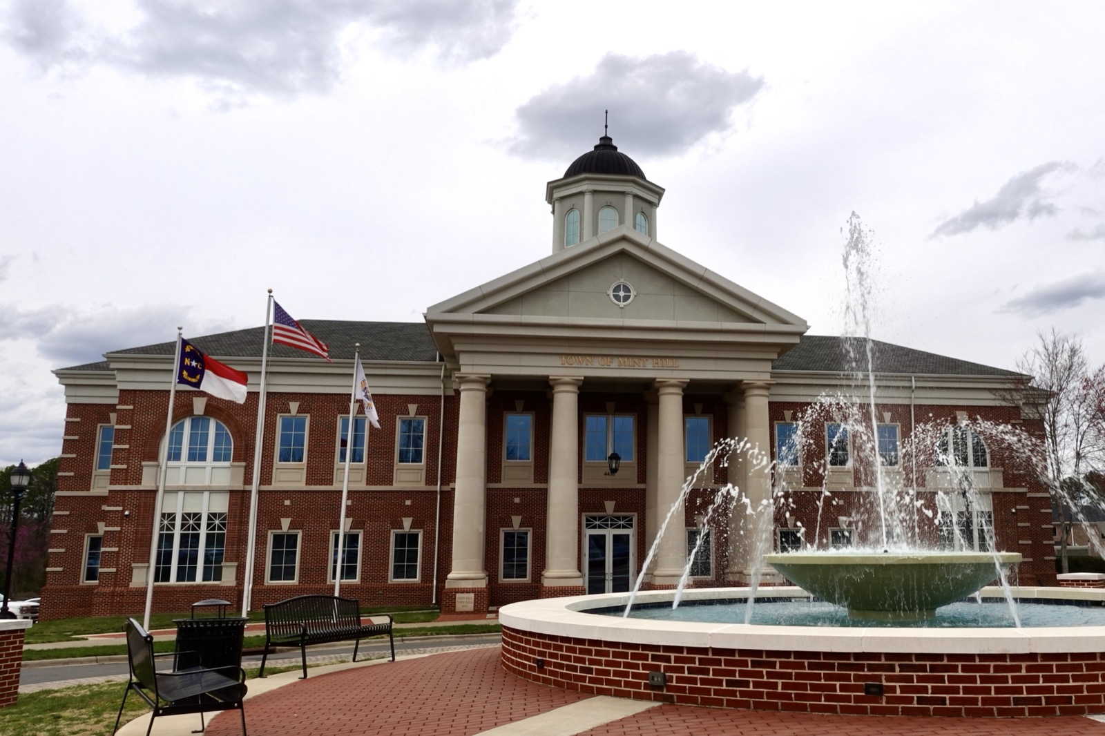 Red brick Mint Hill Town Hall with white columns, a cupola and TOWN OF MINT HILL carved above the entrance, with North Carolina and United States flags and a tiered fountain in the foreground
